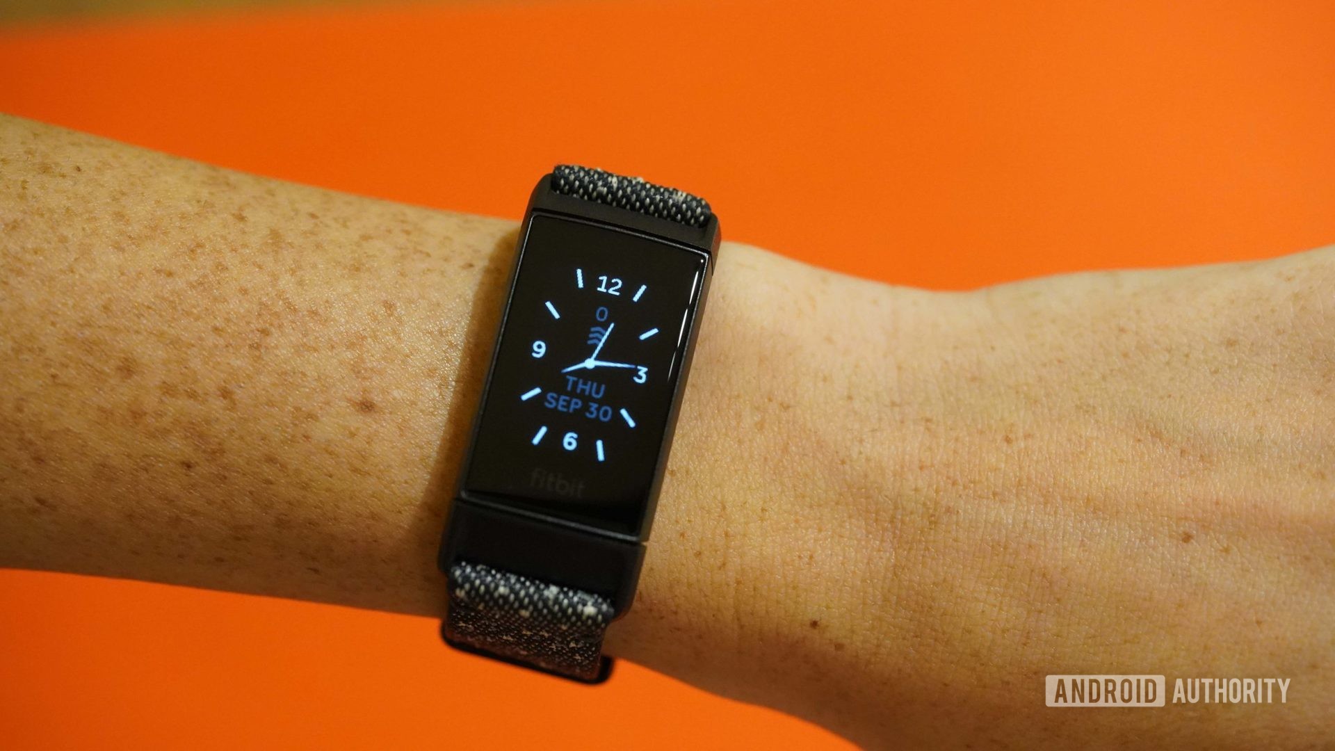 A Fitbit Charge 4 displays the a clock face against an orange background