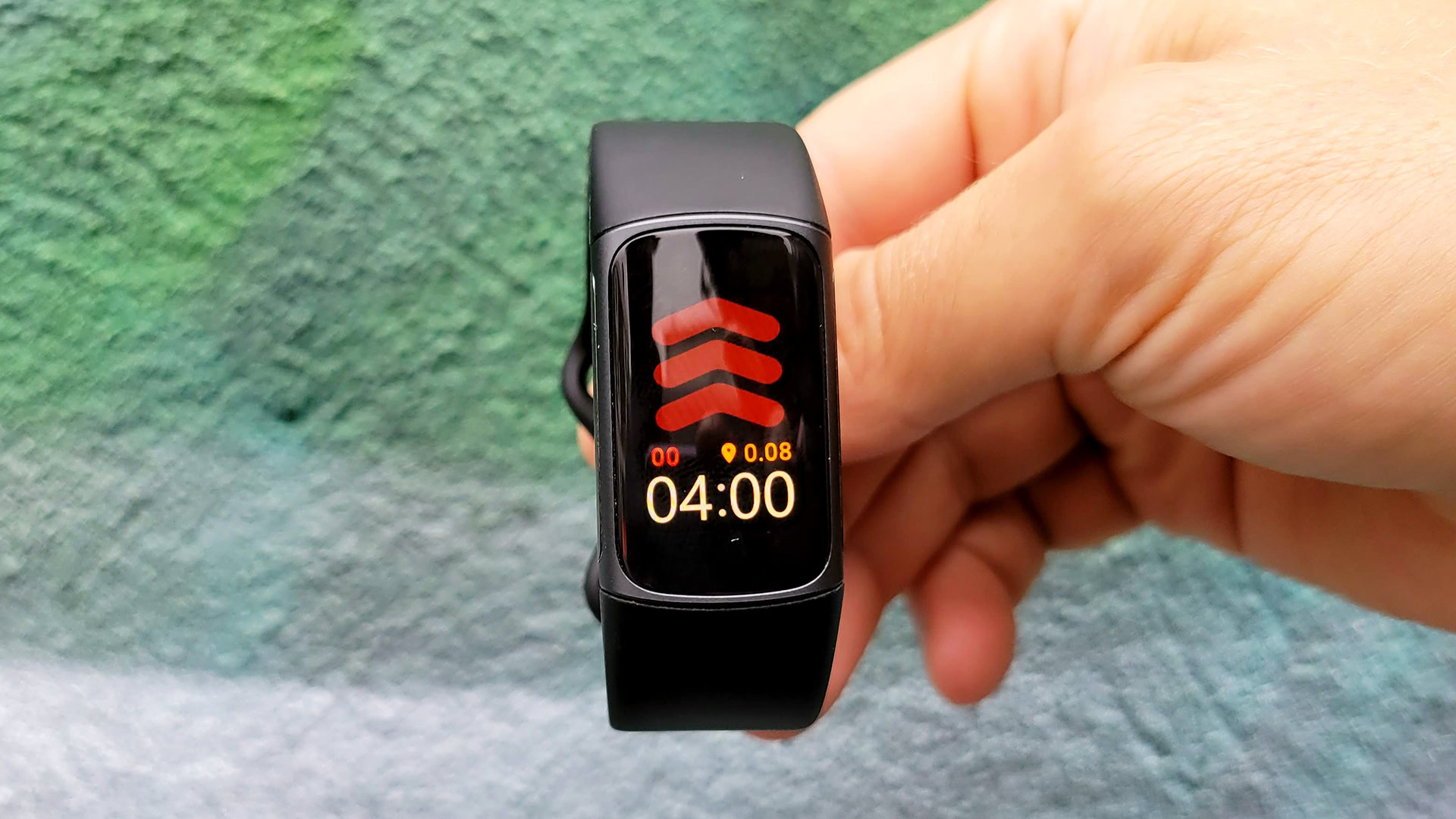 A user holds up a Fitbit Charge 5 in front of a cement wall painted green.