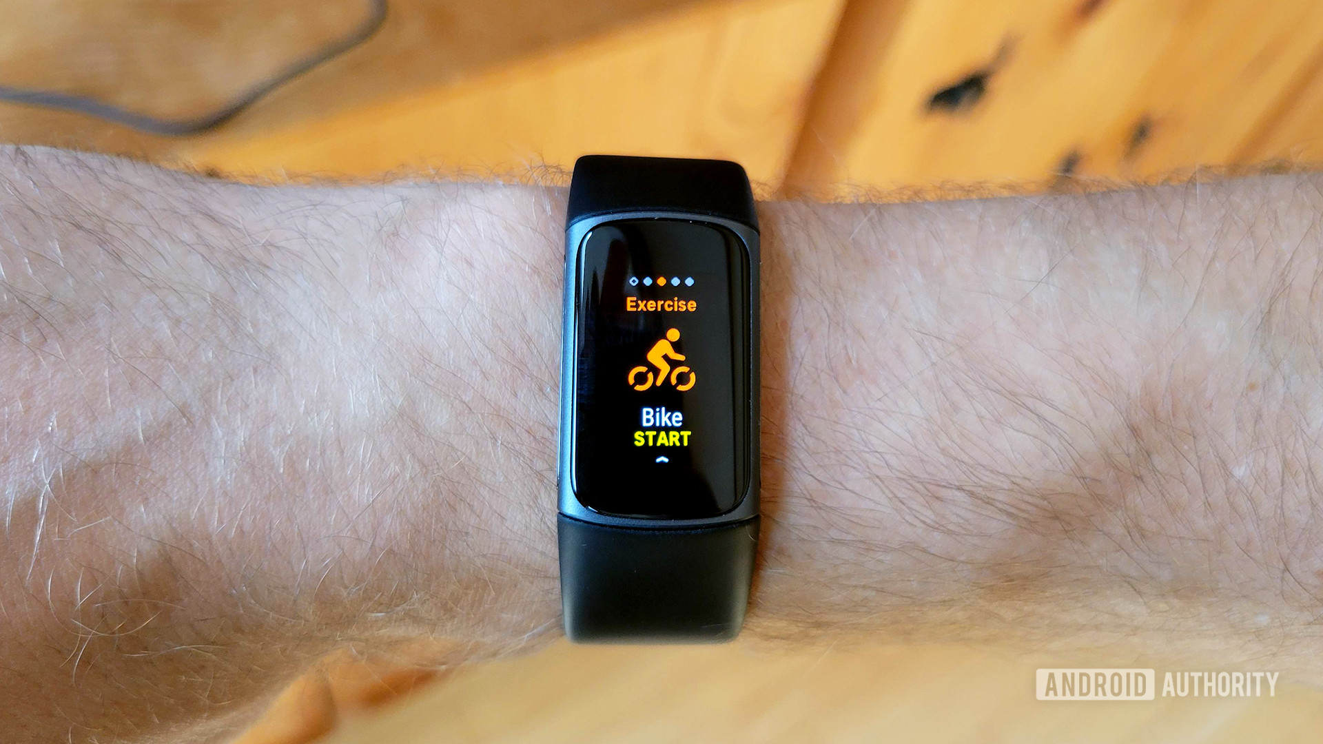 A Fitbit Charge 5 on a user's wrist display the start screen a bike exercise.