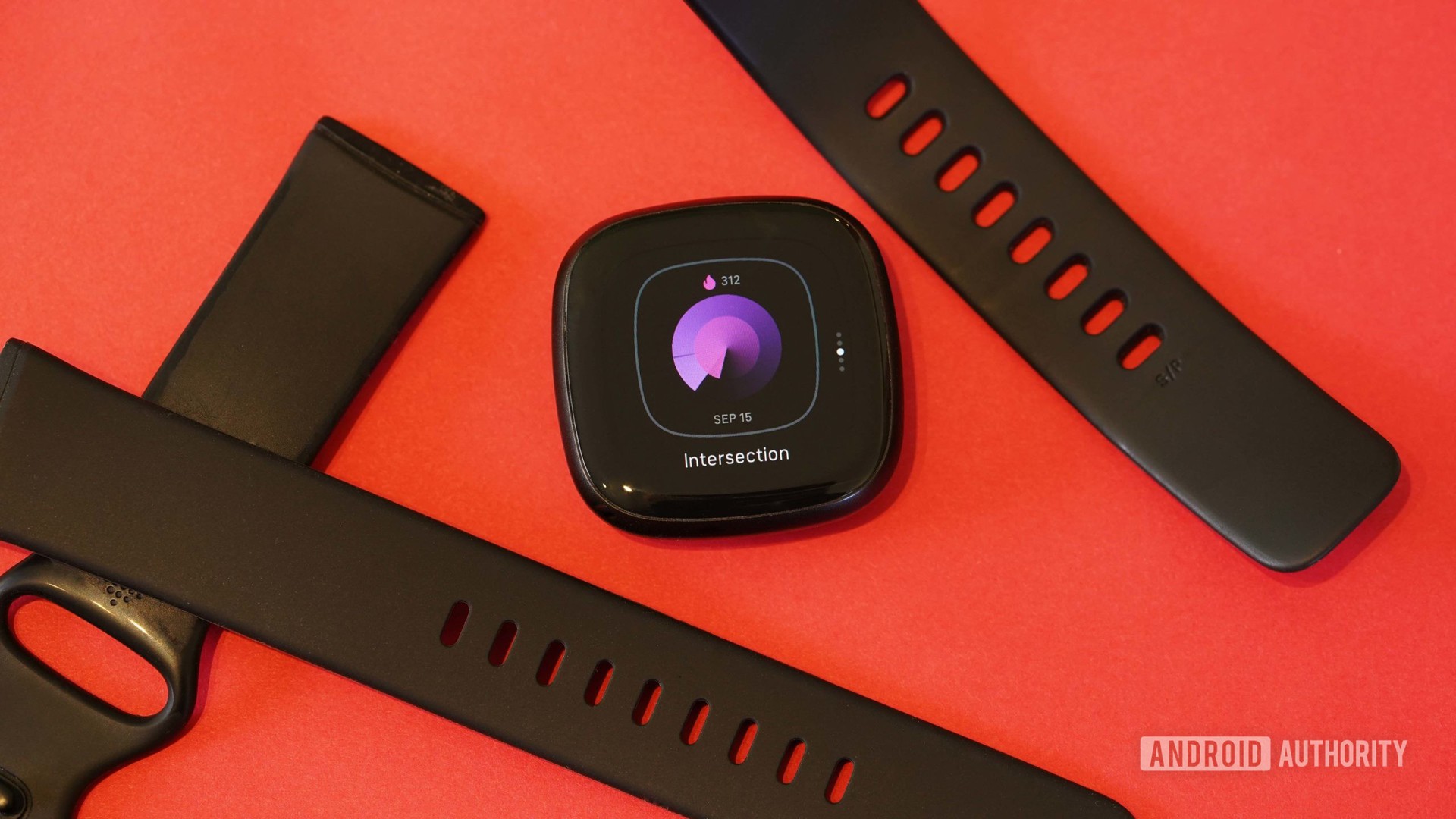 Bir Fitbit Versa 3, bantları ve saat kadranıyla orta değişimde kırmızı bir yüzey üzerinde duruyor.