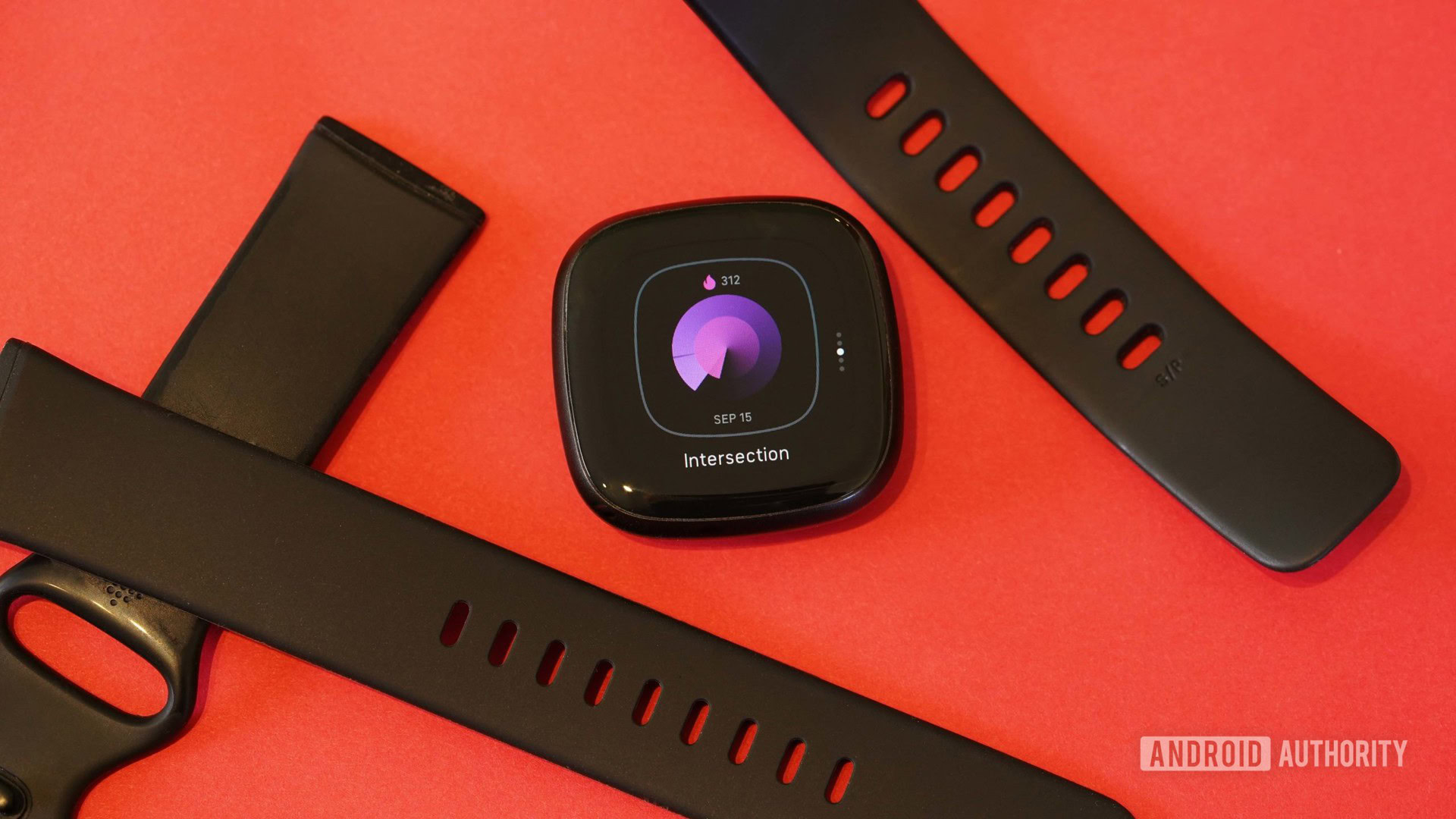 Un Fitbit Versa 3 descansa sobre una superficie roja con las correas y la esfera del reloj cambiando.