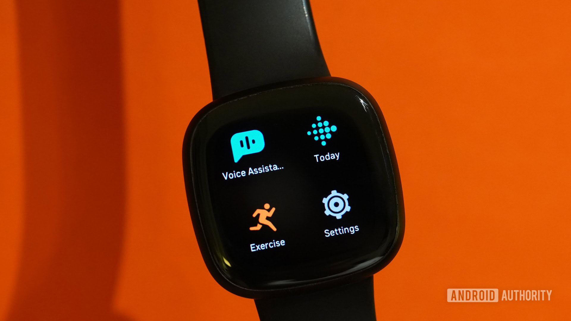 A Fitbit Versa 3 displays apps against an orange background.