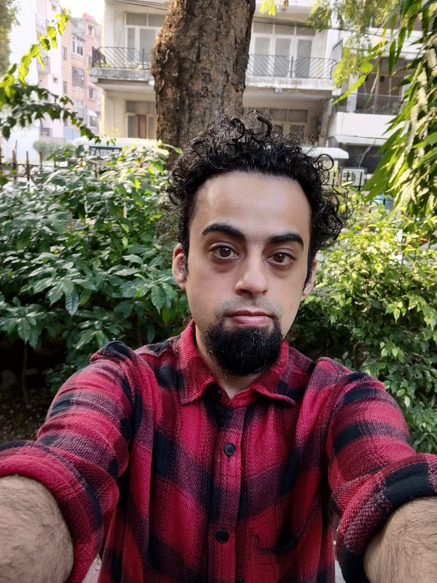 Moto Edge 20 pro selfie camera of a man with dark curly hair and a beard, wearing a red and black checked shirt and standing in front of green bushes.