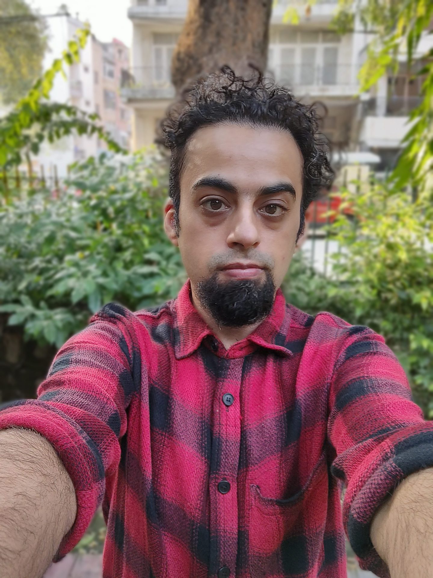 Moto Edge 20 pro selfie portrait of a man with dark curly hair and a beard, wearing a red and black checked shirt and standing in front of green bushes.