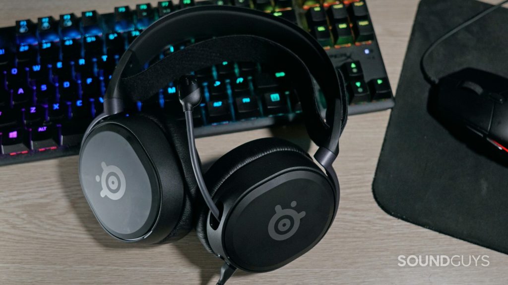 The SteelSeries Arctis Prim gaming headset lays on a desk next to a HyperX mechanical gaming keyboard and a Logitech gaming mouse.