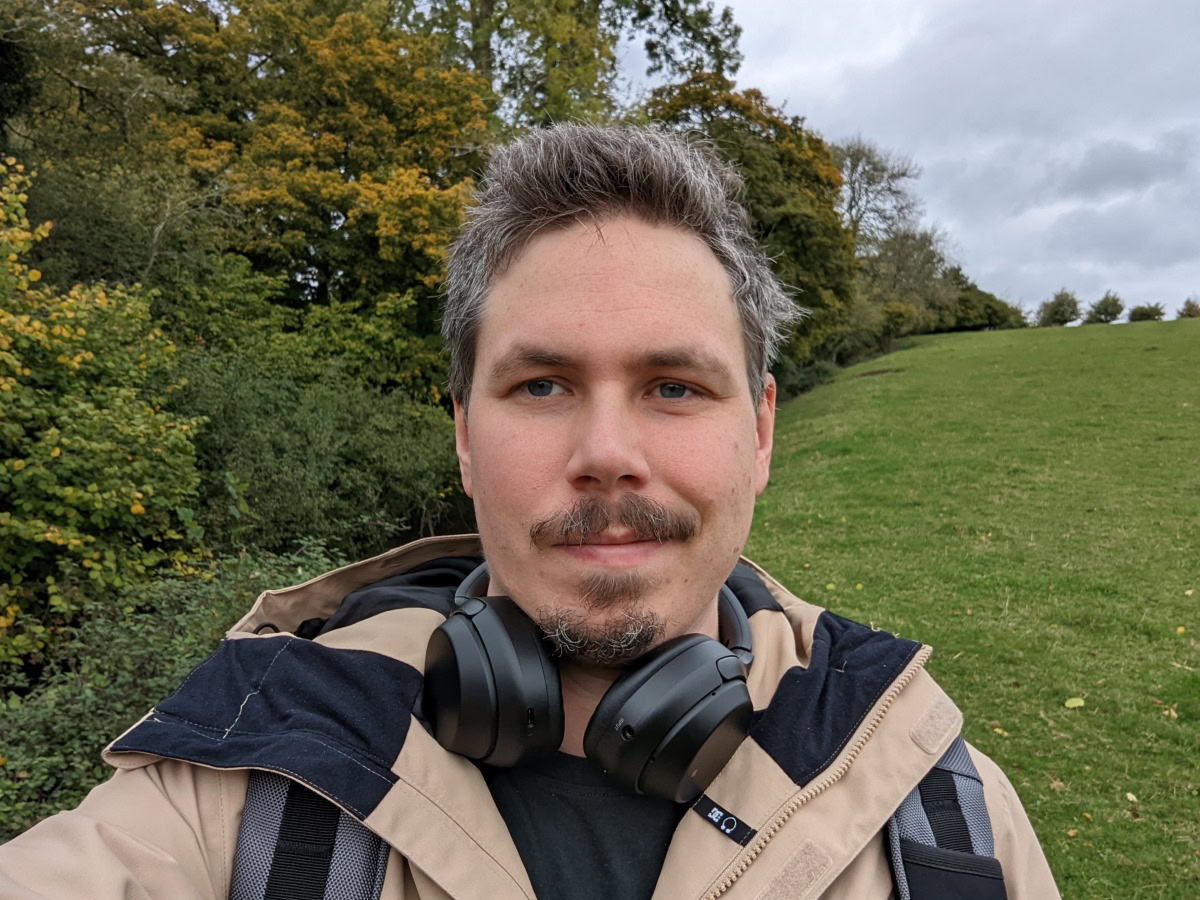 Camera sample selfie outdoor shot of a man with dark hair and facial hair wearing a beige jacket, with headphones around his neck, taken on the Google Pixel 6 Pro