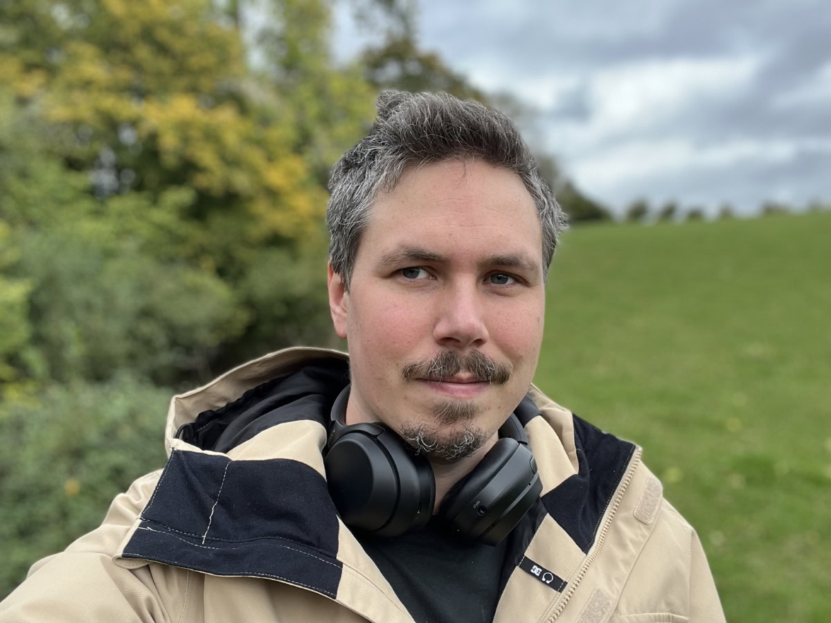 Camera sample selfie outdoor portrait shot of a man with dark hair and facial hair, wearing a beige jacket, with headphones around his neck, taken on the Apple iPhone 13 Pro Max
