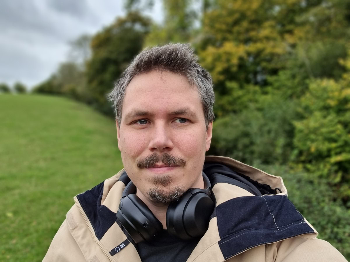 Camera sample selfie outdoor portrait shot of a man with dark hair and facial hair, wearing a beige jacket, with headphones around his neck, taken on the Samsung Galaxy S21 Ultra