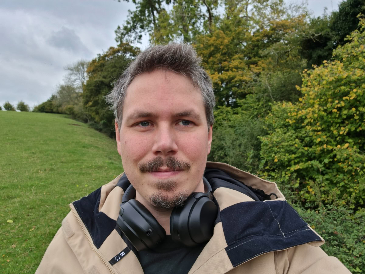Camera sample selfie outdoor shot of a man with dark hair and facial hair, wearing a beige jacket, with headphones around his neck, taken on the Samsung Galaxy S21 Ultra