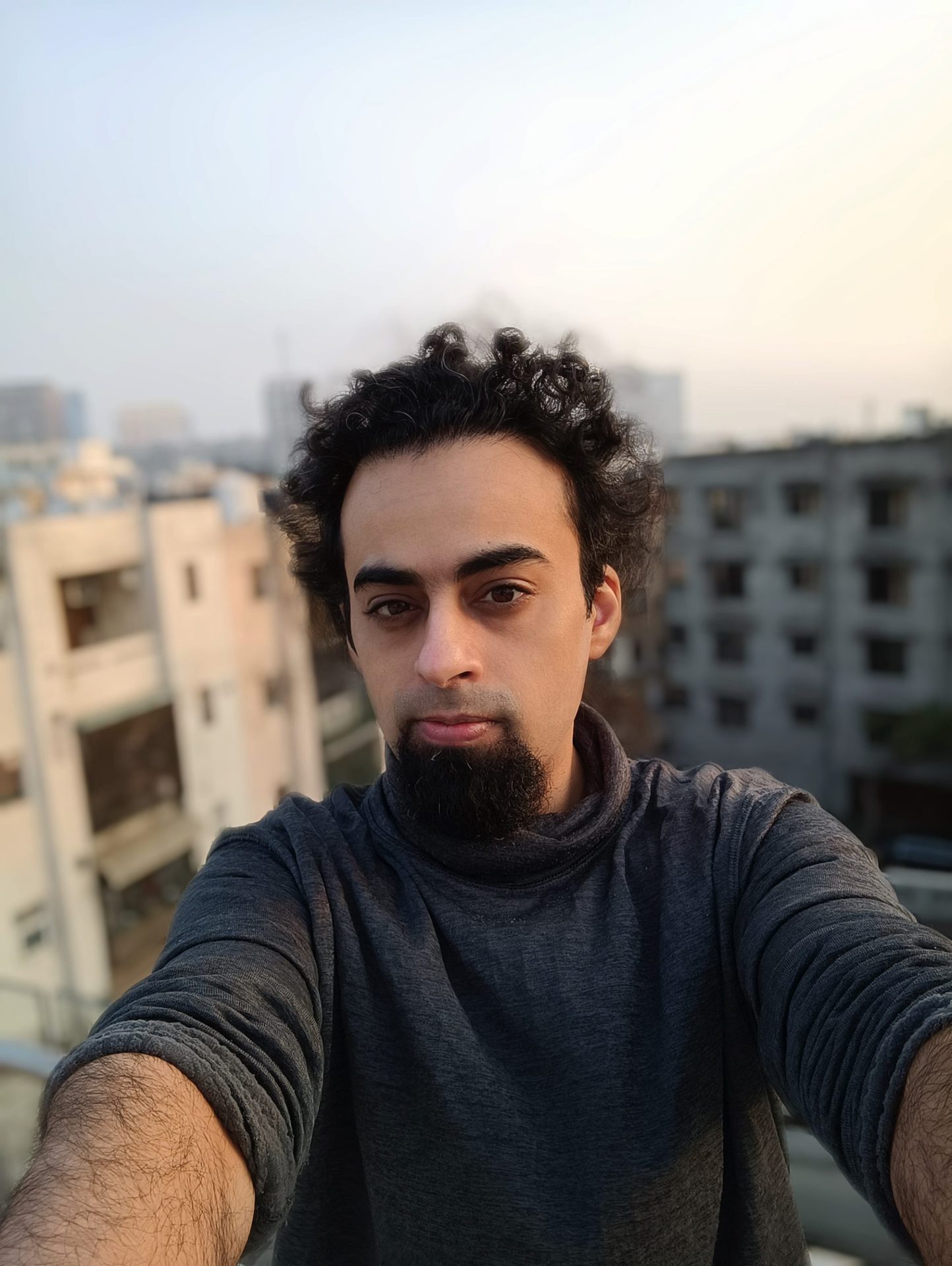 Mi 11i front camera portrait mode of a man with dark curly hair and a beard, with buildings visible behind him