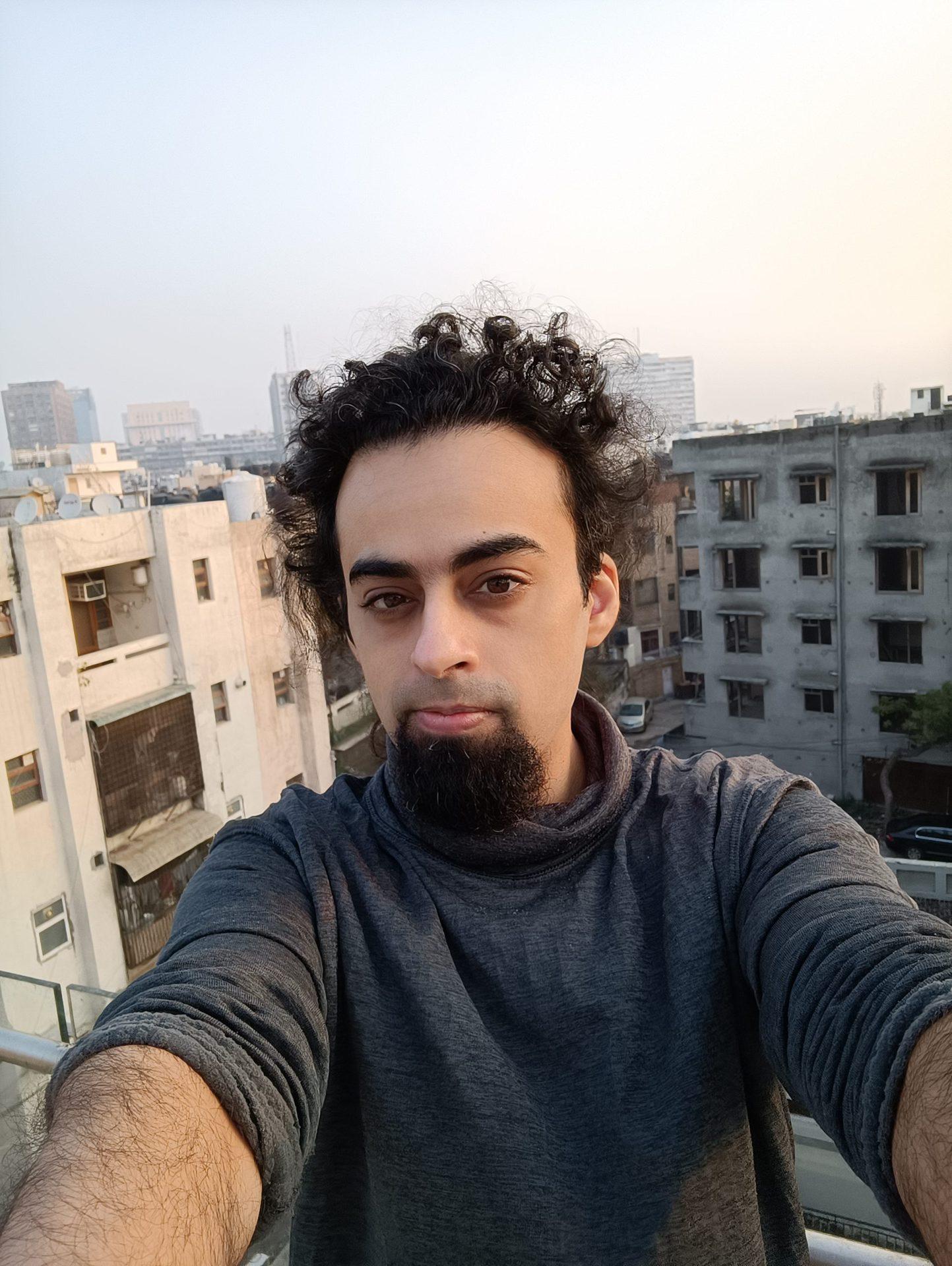 Mi 11i front camera selfie of a man with dark curly hair and a beard, with buildings visible behind him