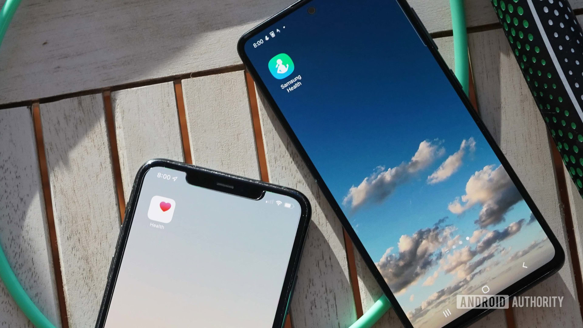 A Samsung Galaxy A51 and Apple iPhone 11 rest on a wooden table, each displaying their respective health app.