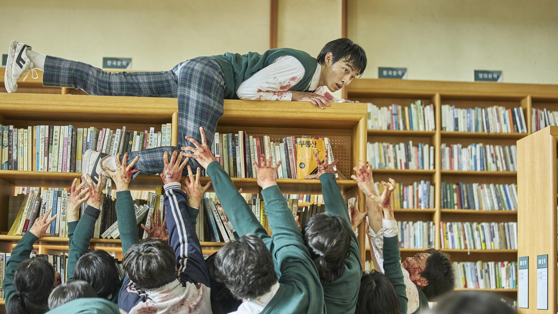 A still from All of Us Are Dead on Netflix showing a school pupil on top of a bookcase as zombies reach for him.