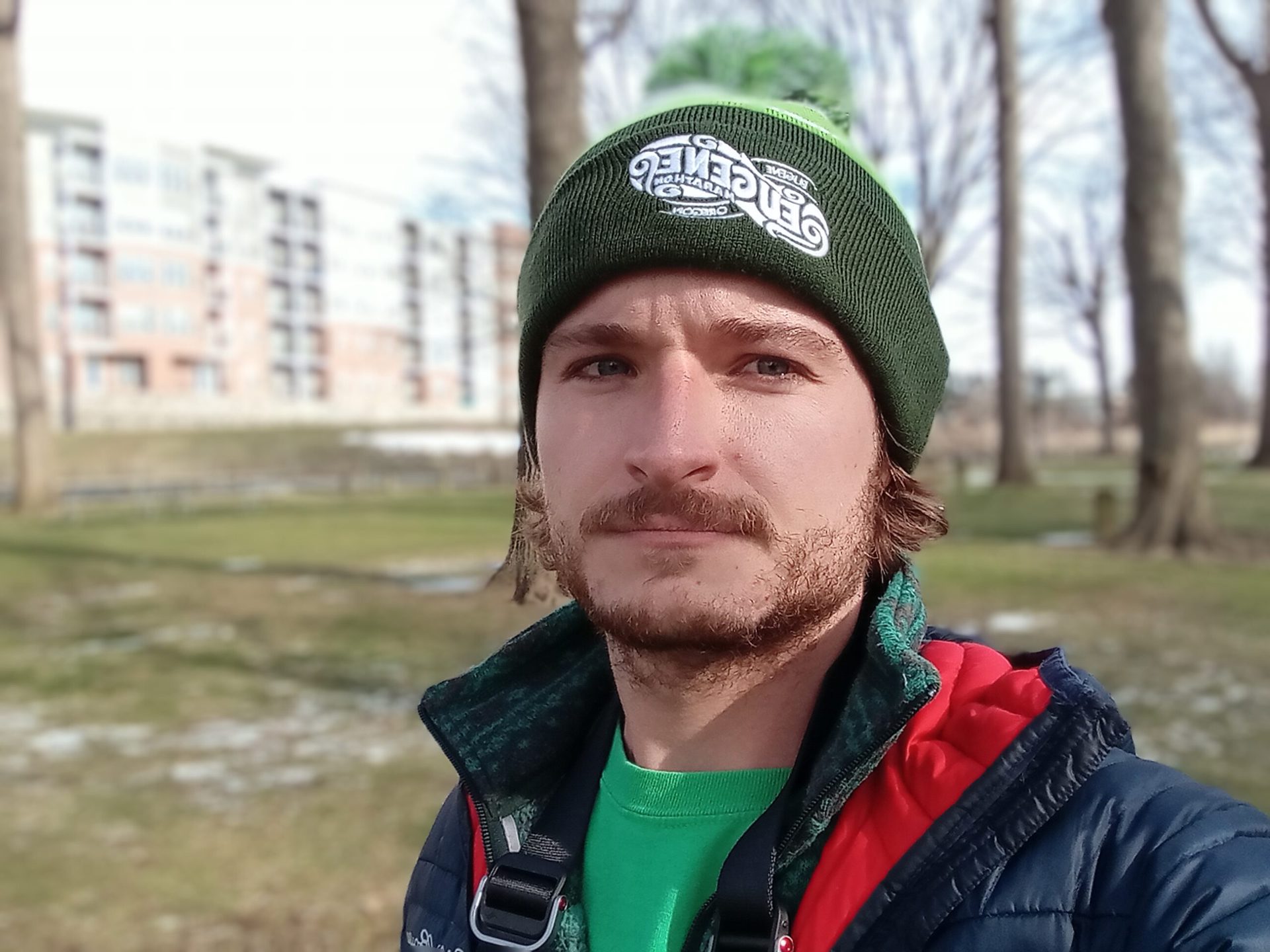galaxy a03s portrait selfie outdoors of a man with facial hair wearing a green hat, green t-shirt, and red and black coat, with buildings and trees visible behind him.