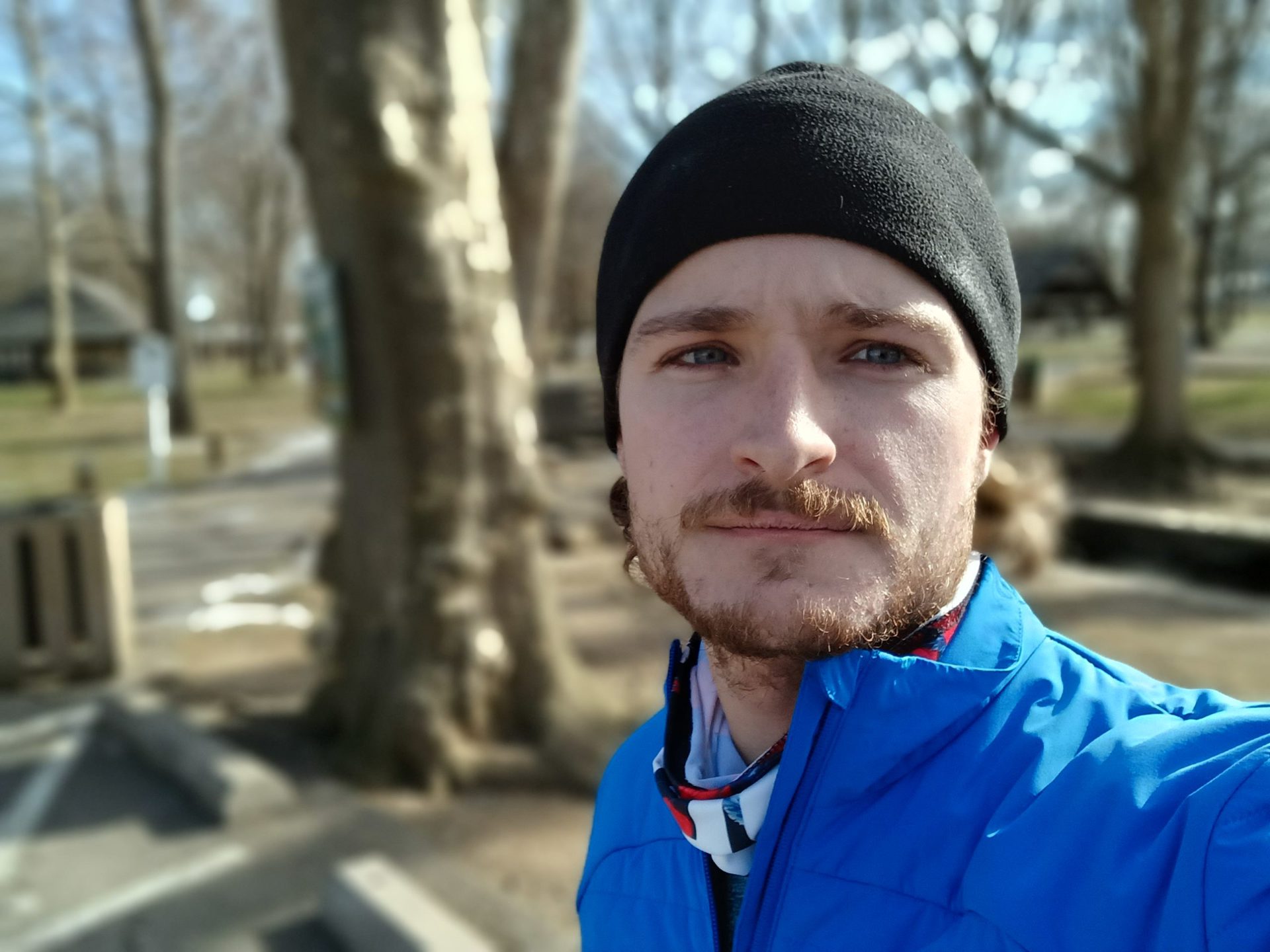 Galaxy A13 portrait selfie taken outdoors of a man with facial hair wearing a black hat and a bright blue jacket