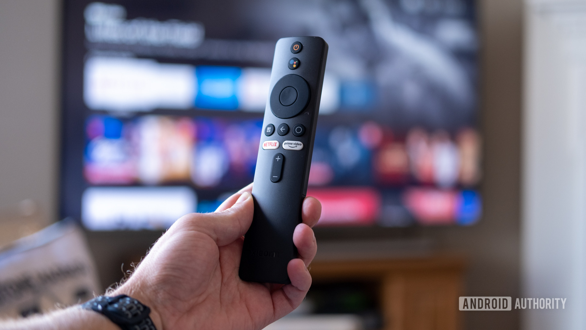 Xiaomi TV Stick remote in hand, in front of TV.