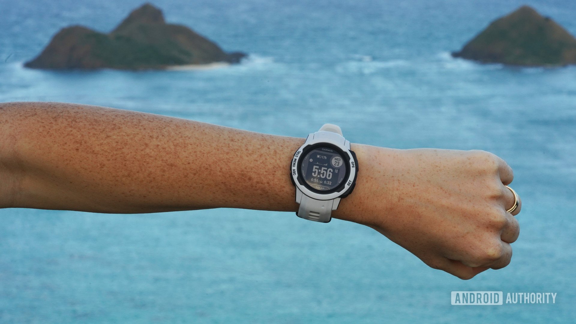 A user models the Garmin Instinct 2S in Mist Gray at the peak of an ocean front hike.