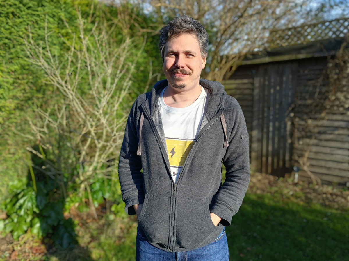 Huawei P50 Pro camera portrait of man with brown hair and facial hair wearing a white t-shirt with a yellow logo, a grey hoodie, and blue jeans in front of hedge