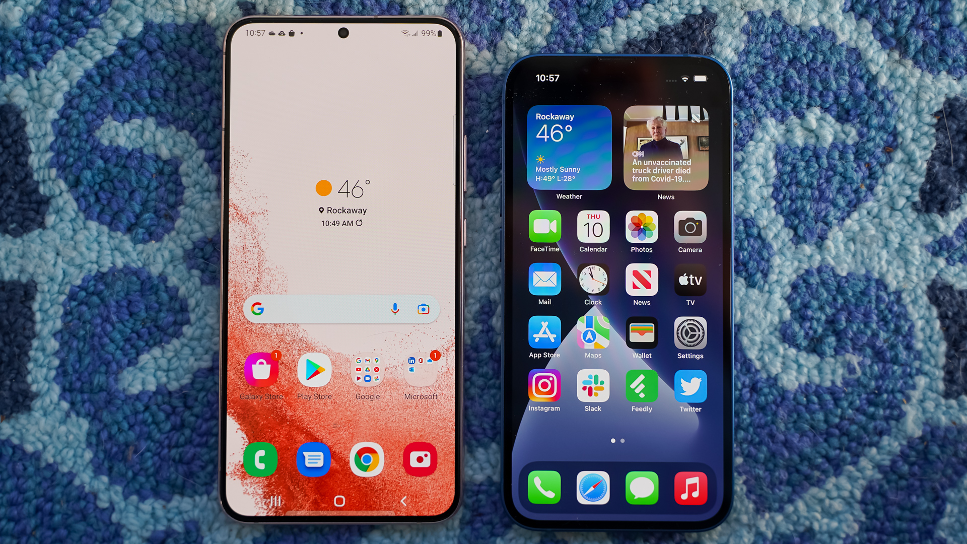 Un Samsung Galaxy S22 Plus junto a un Apple iPhone 13 mostrando sus pantallas de inicio.  Ambos descansan sobre una alfombra azul estampada.