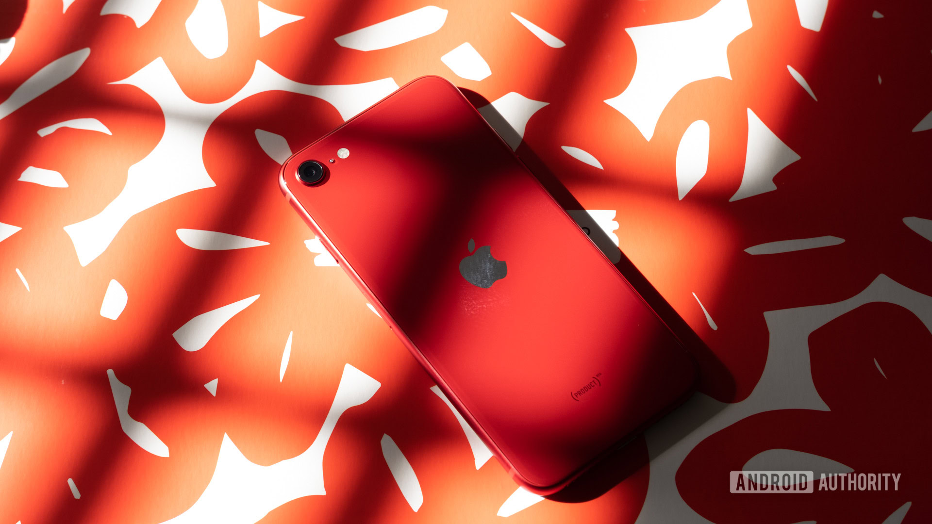iPhone SE face down showing rear with camera and Apple logo, on a white and red patterned background.