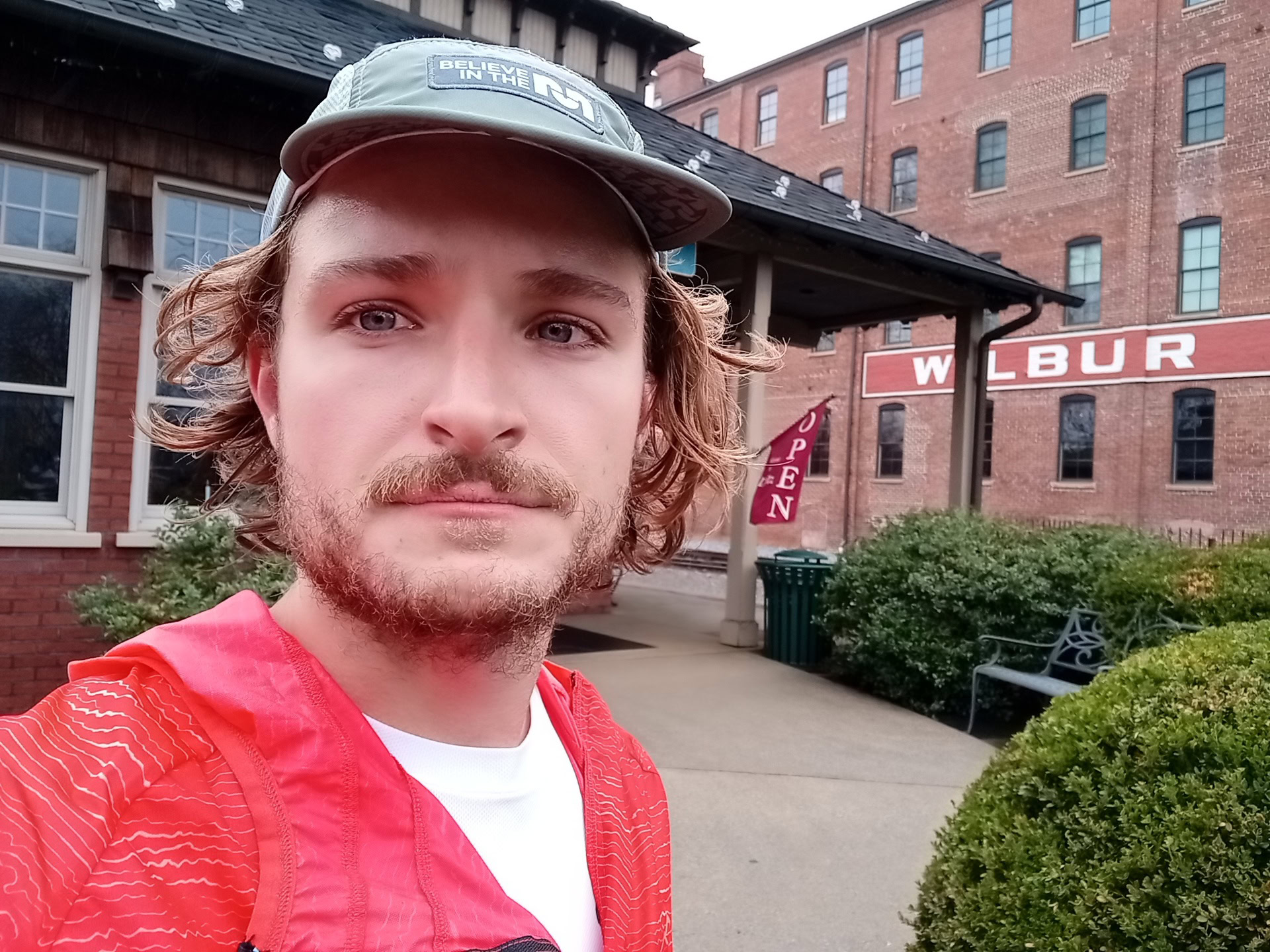 Moto G Stylus beauty filter selfie of a man with fair curly hair and facial hair wearing a white t-shirt and red top, with a baseball cap, outdoors in front of a red brick building.