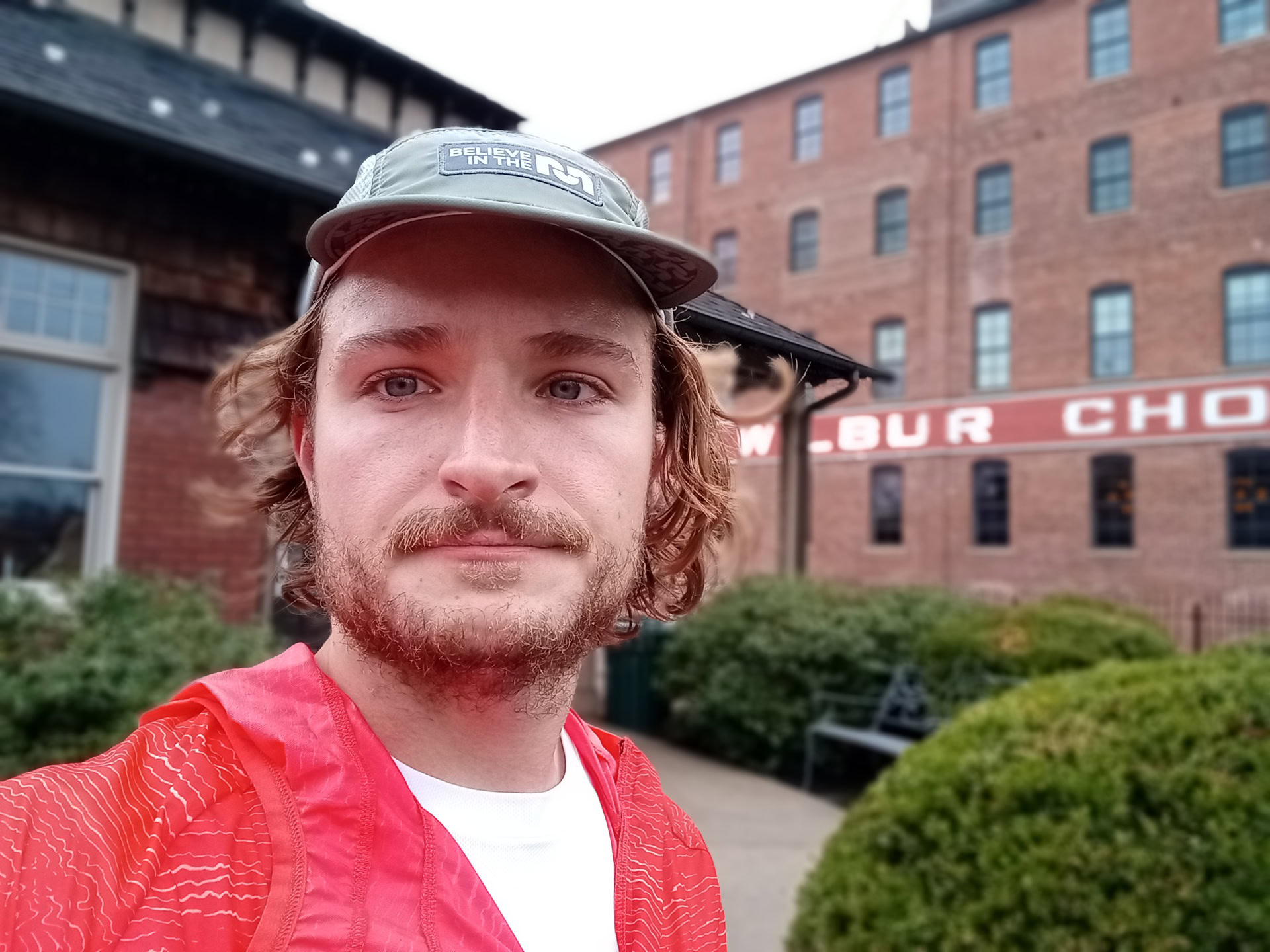 Moto g stylus portrait selfie fair curly hair and facial hair wearing a white t-shirt and red top, with a baseball cap, outdoors in front of a red brick building.