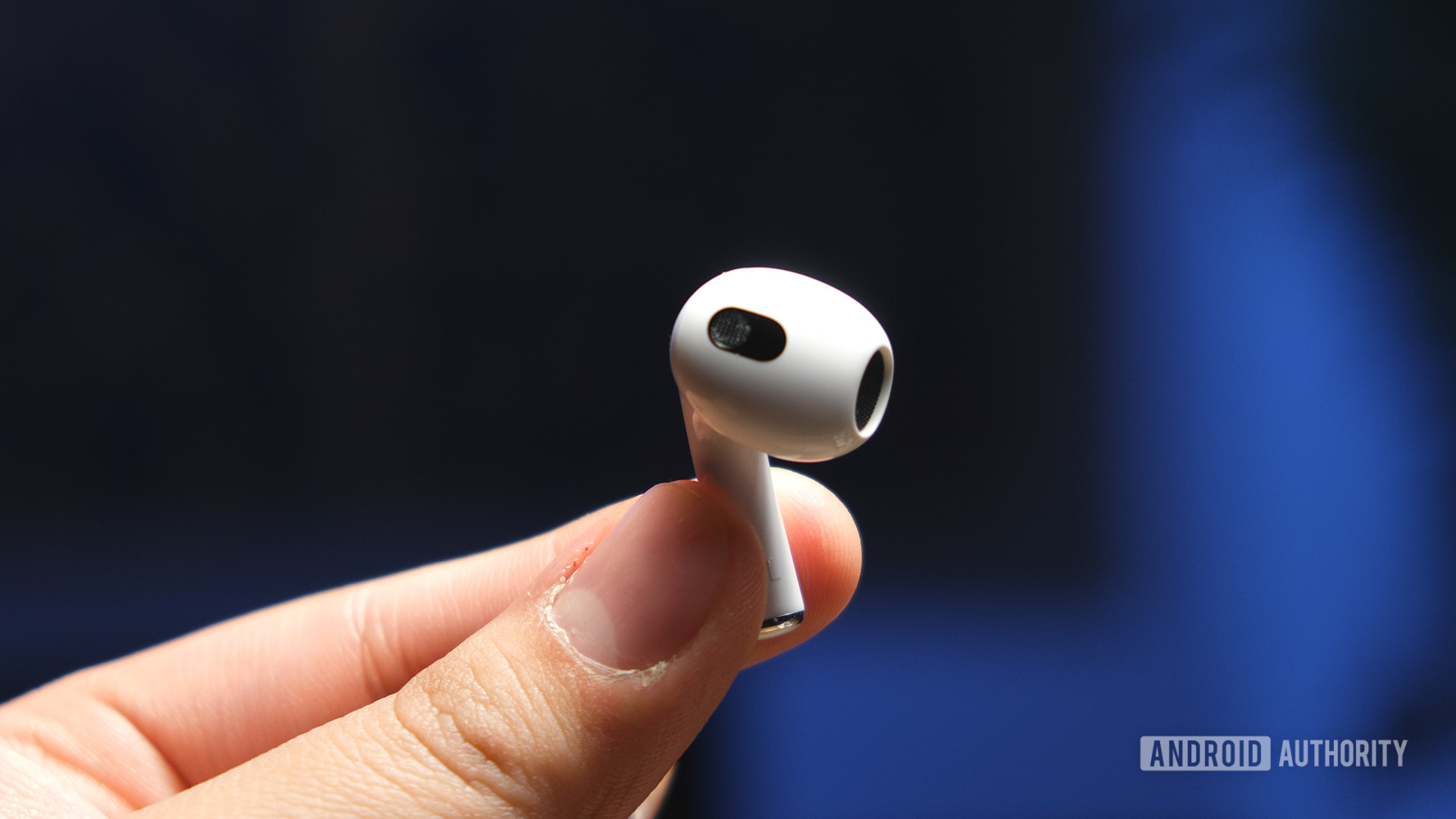 A hand holds a Apple AirPods (3rd generation) earbud by the stem to reveal the open-type fit and embedded sensors.
