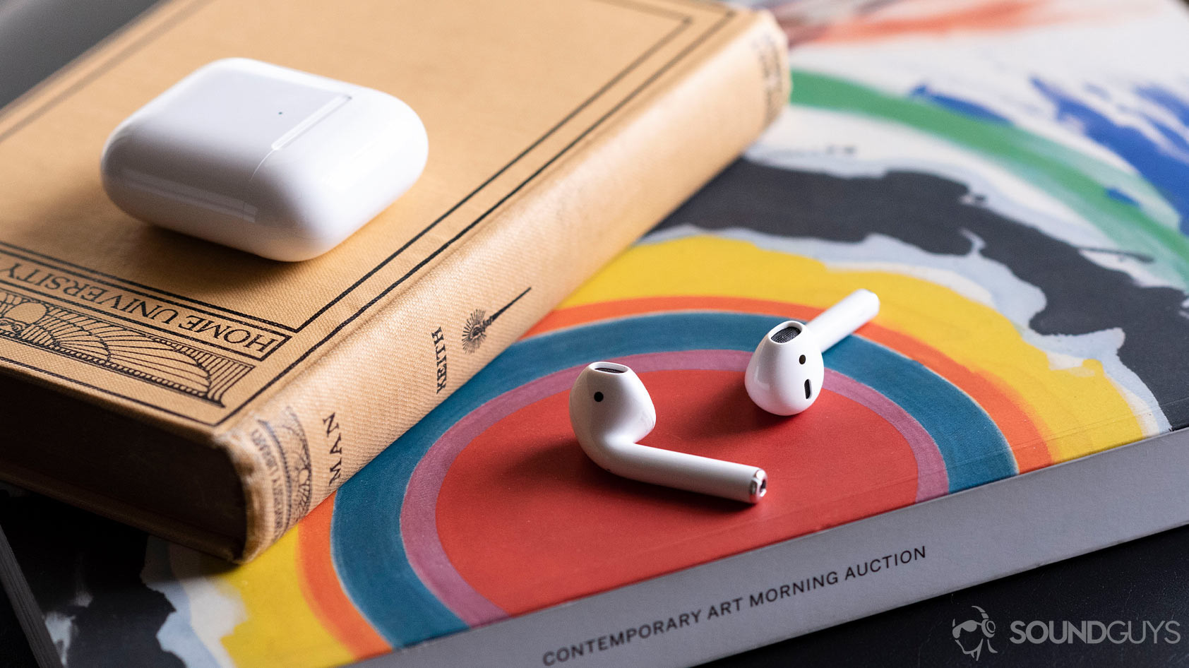 The Apple AirPods (2nd generation) on an arts magazine with the case above it, shut.