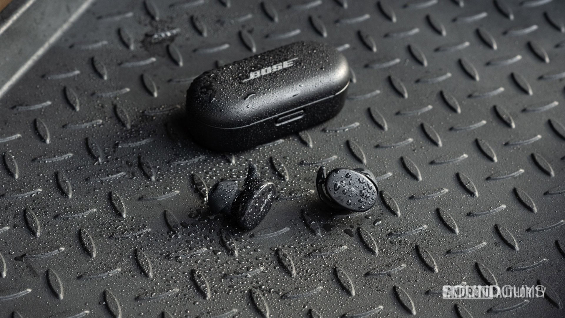 The Bose Sport Earbuds true wireless workout earbuds sit outside of the closed charging case, all objects are covered in sprinkles of water.