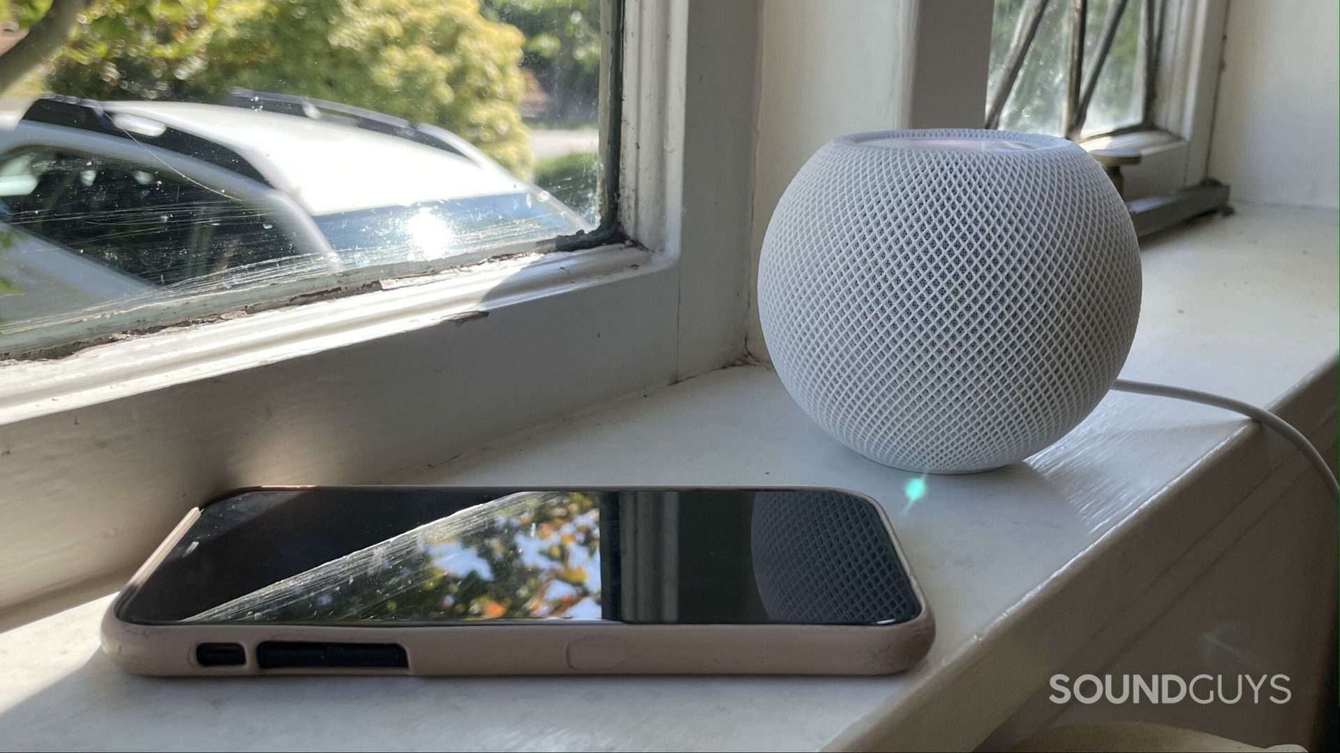 The Apple HomePod mini on a window sill next to an iPhone.