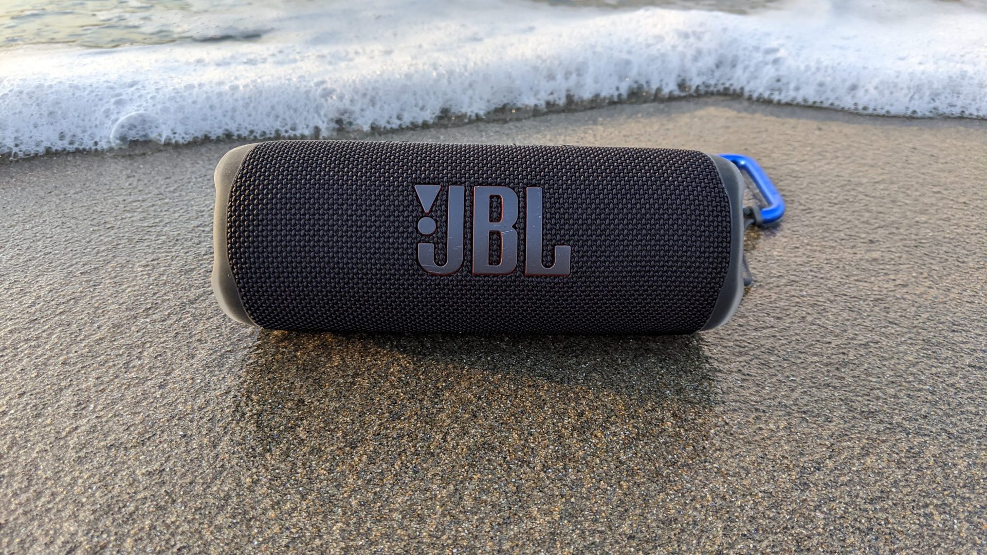 A JBL Flip 6 Bluetooth speaker sitting on wet sand by the ocean.
