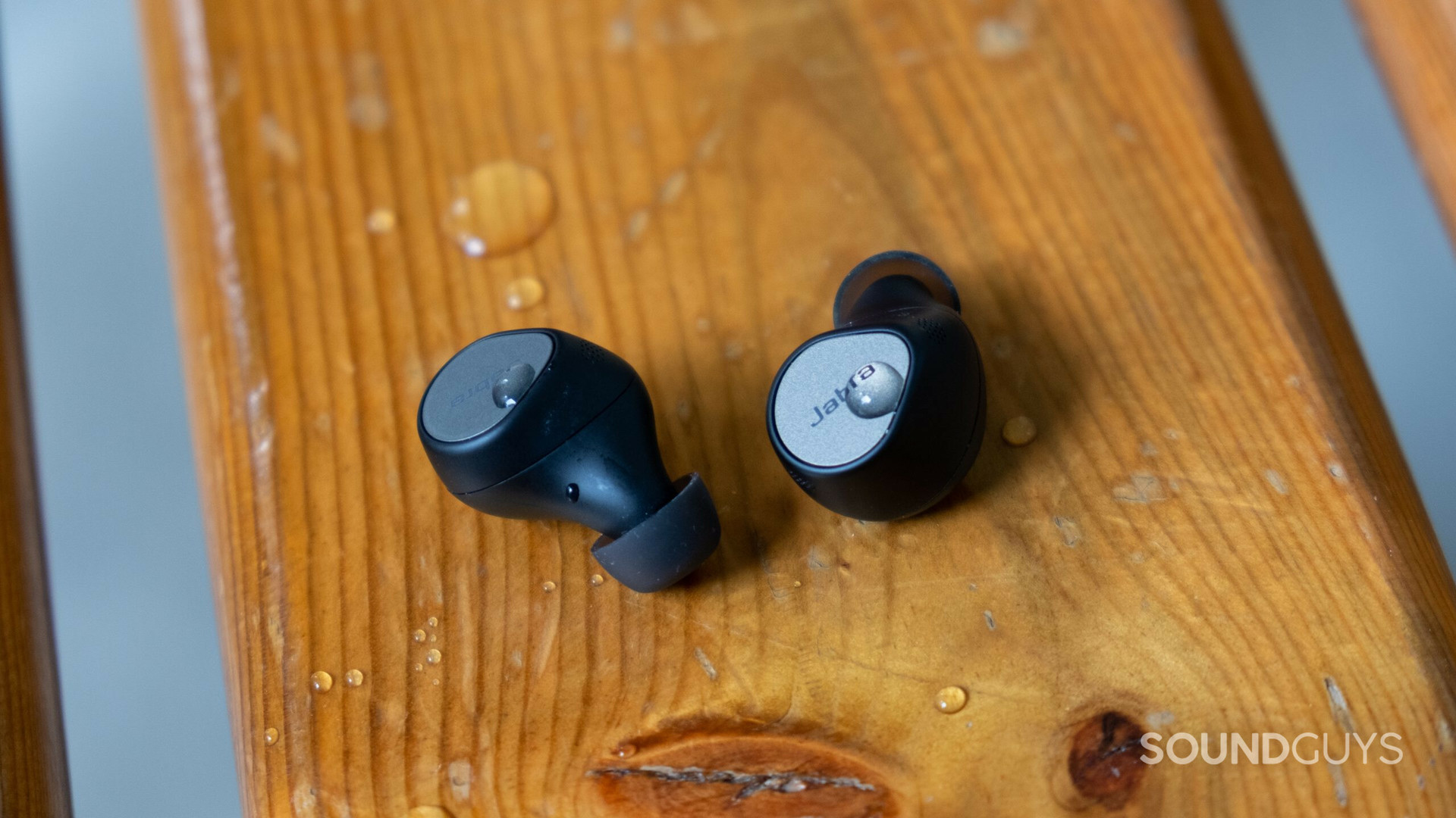 The Jabra Elite 7 Pro earbuds sitting on a bench with some water dropson the buds and the bench.