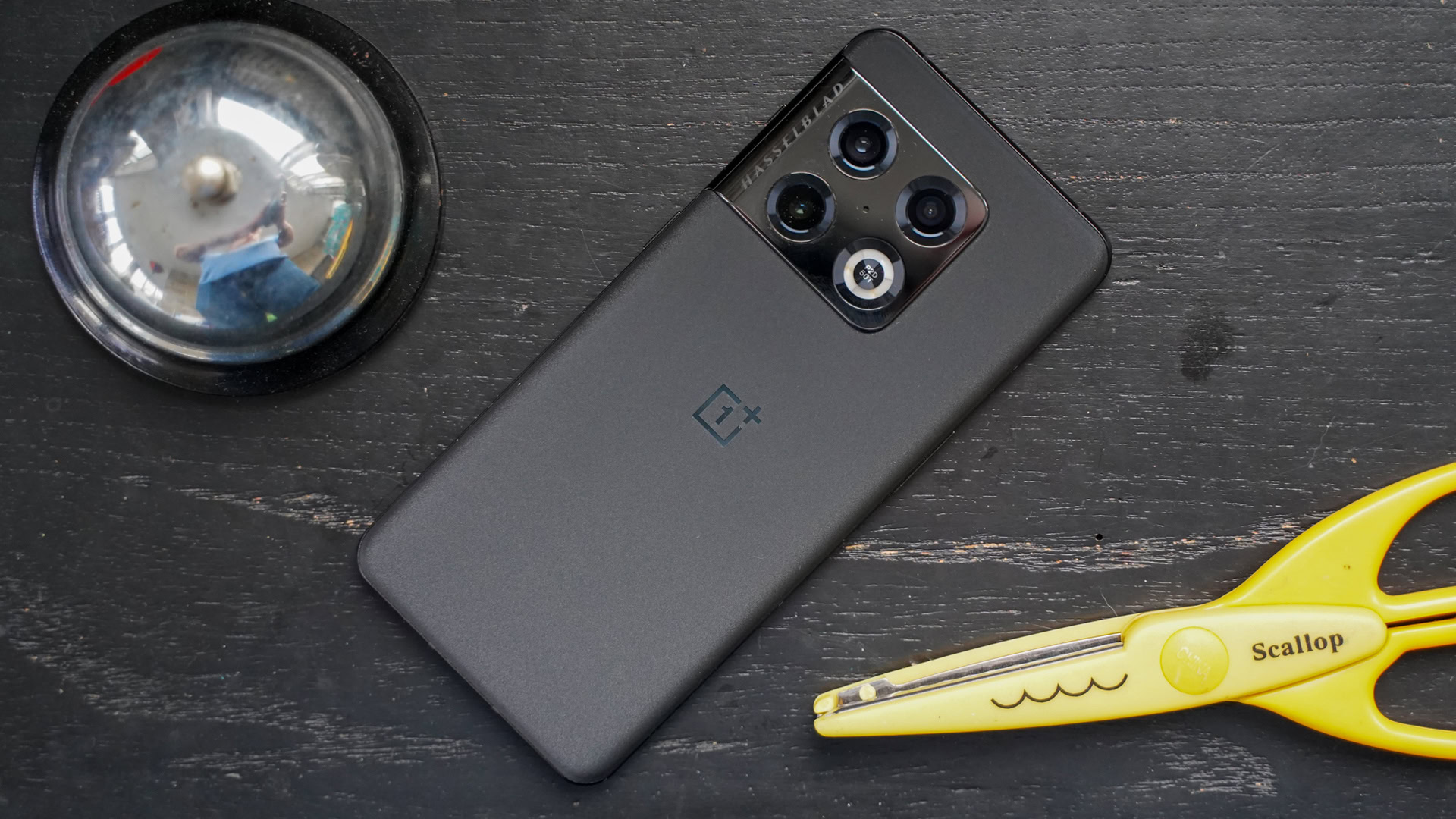 OnePlus 10 Pro rear view showing cameras, next to a bell and a pair of yellow scissors