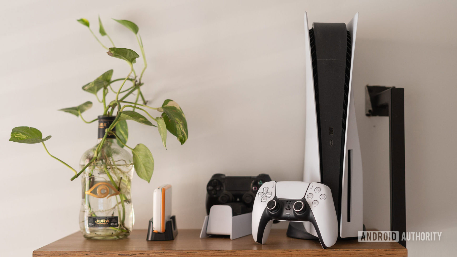 A PlayStation 5 next to two controllers sitting on a table near a vase with a plant in it.