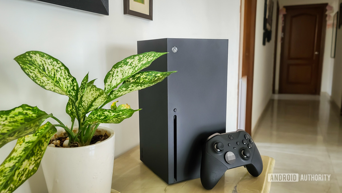 An Xbox Series X sitting next to a controller near a potted plant.
