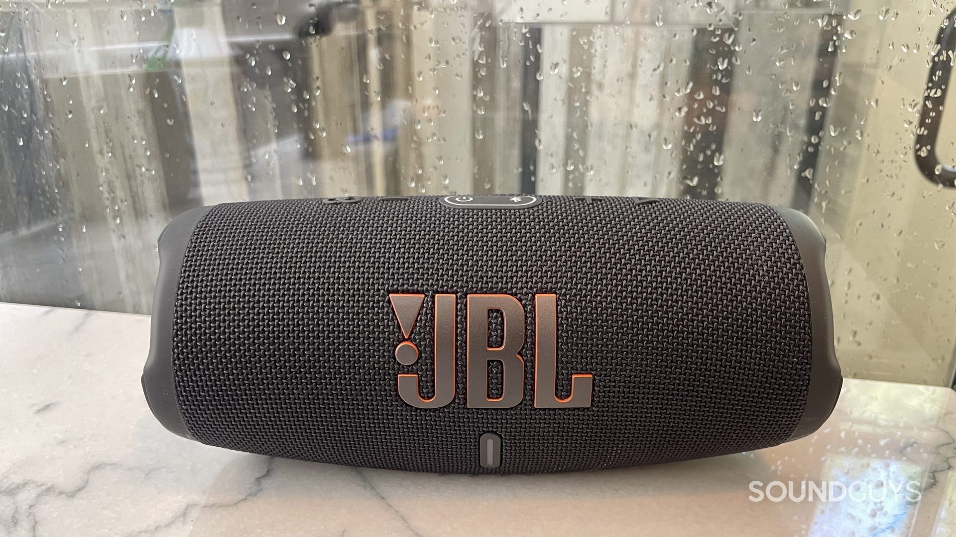 The JBL Charge 5 Bluetooth speaker rests on a countertop with rain drops on a window behind it.