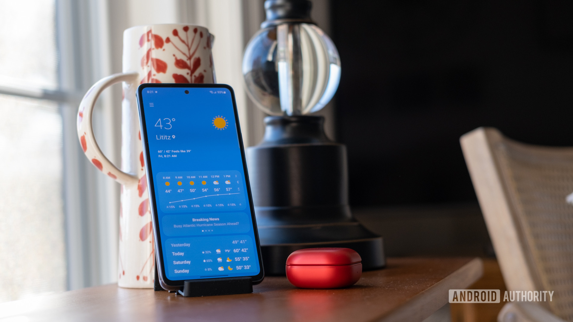 Samsung Galaxy A53 standing upright in a dock, showing weather app, with a red Galaxy Buds case next to it.