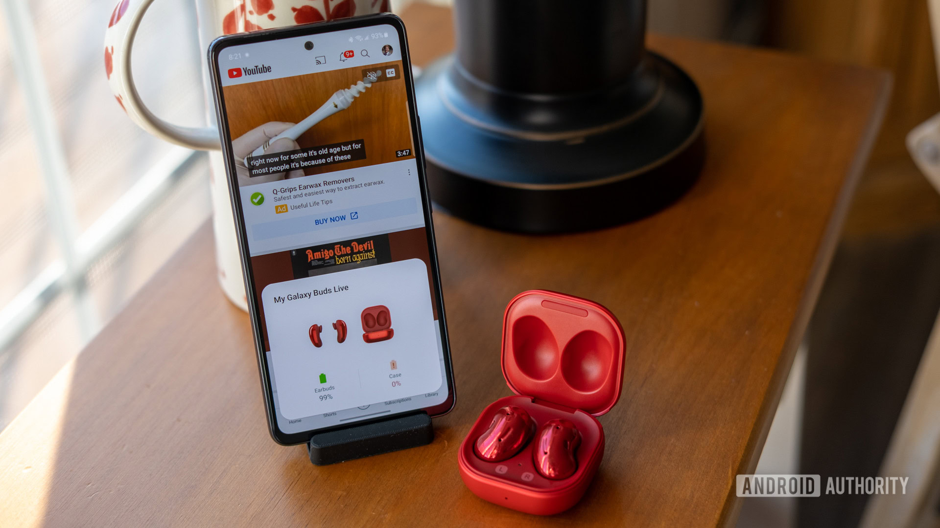 Samsung Galaxy A53 standing upright in a dock showing YouTube on the display, with a pair of red Galaxy Buds in a case next to it.
