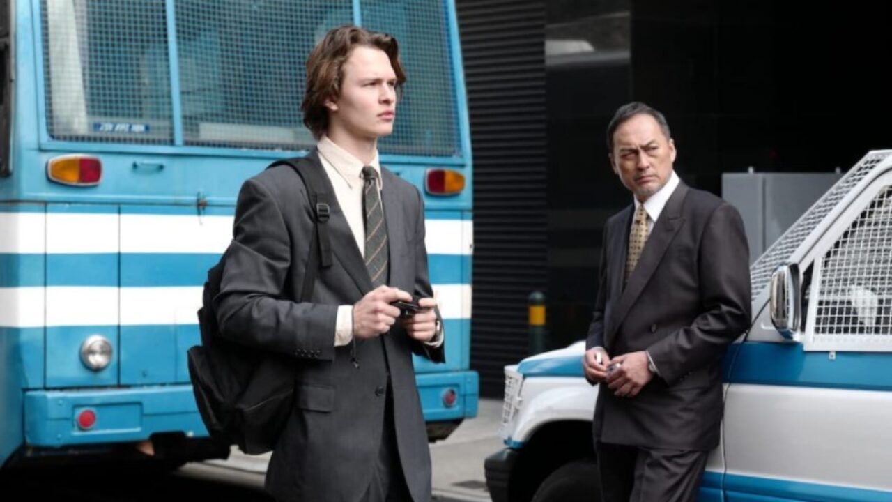 Two Tokyo Vice characters, one standing and one leaning against a vehicle, with a blue and white bus in the background.