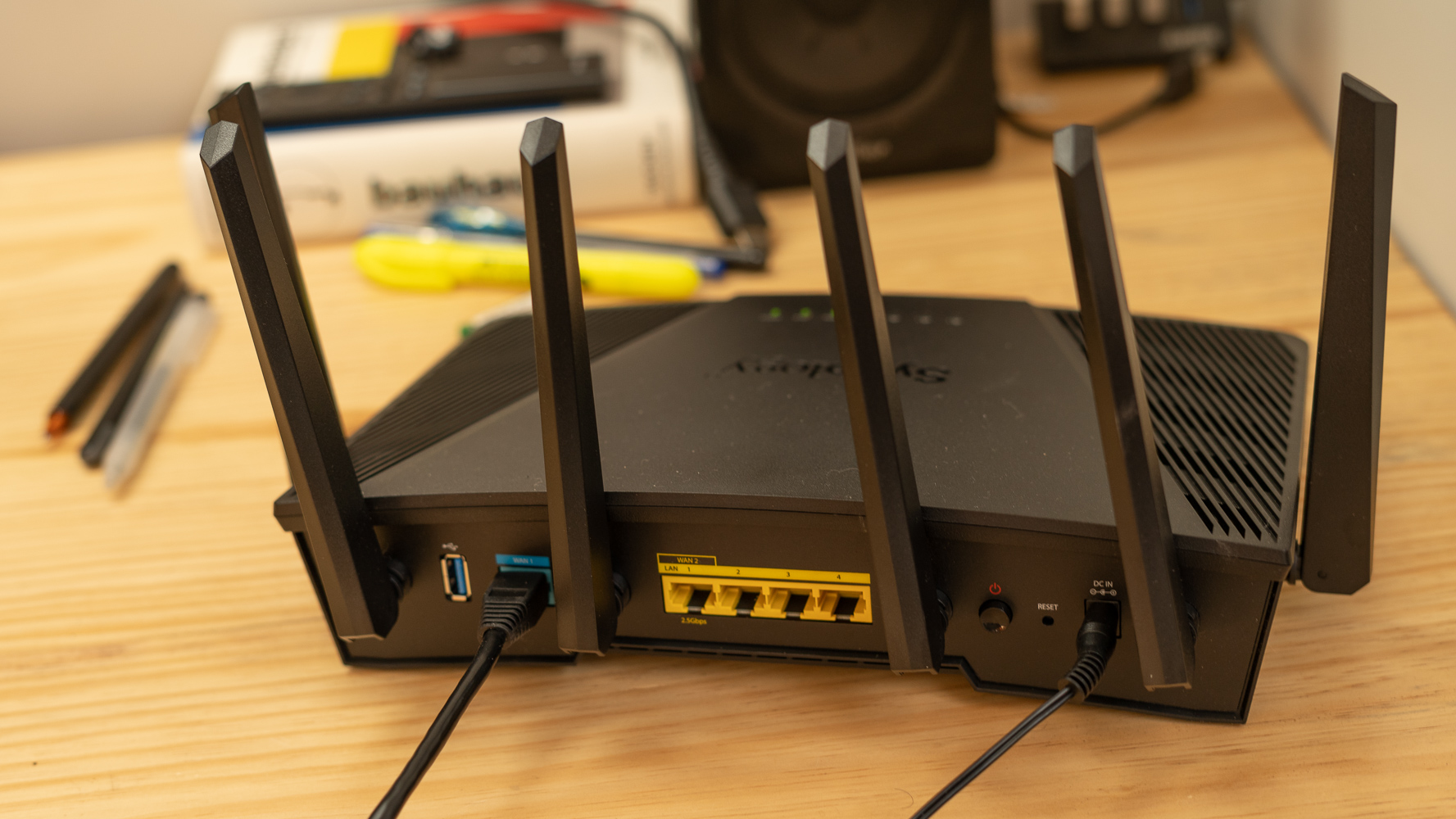 The rear of a Synology RT6600ax Wi-Fi router. It rests on a wooden table in front of some other electornics and books.