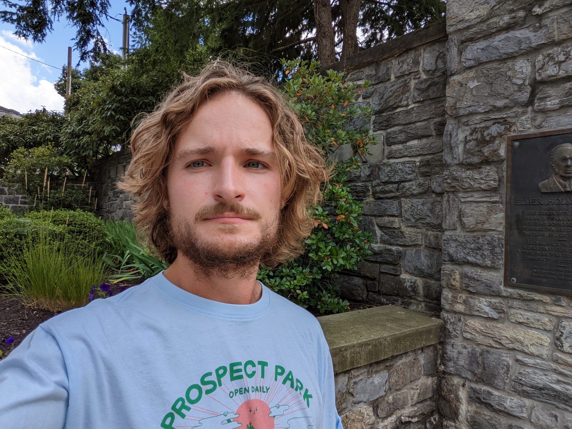 Pixel 6a standard selfie of a man with dirty blonde curly hair in a grey logo T-shirt standing outside with a stone wall visible behind him.