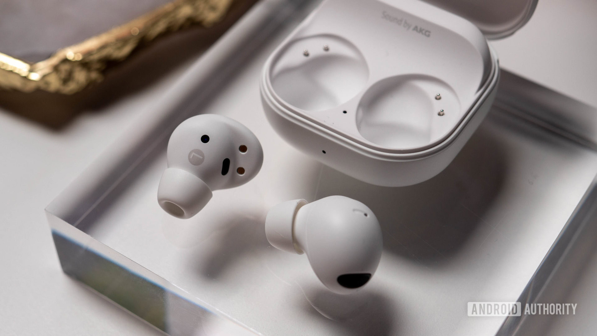 The Samsung Galaxy Buds 2 Pro in white color sitting next to their charging case on a block of clear plastic.