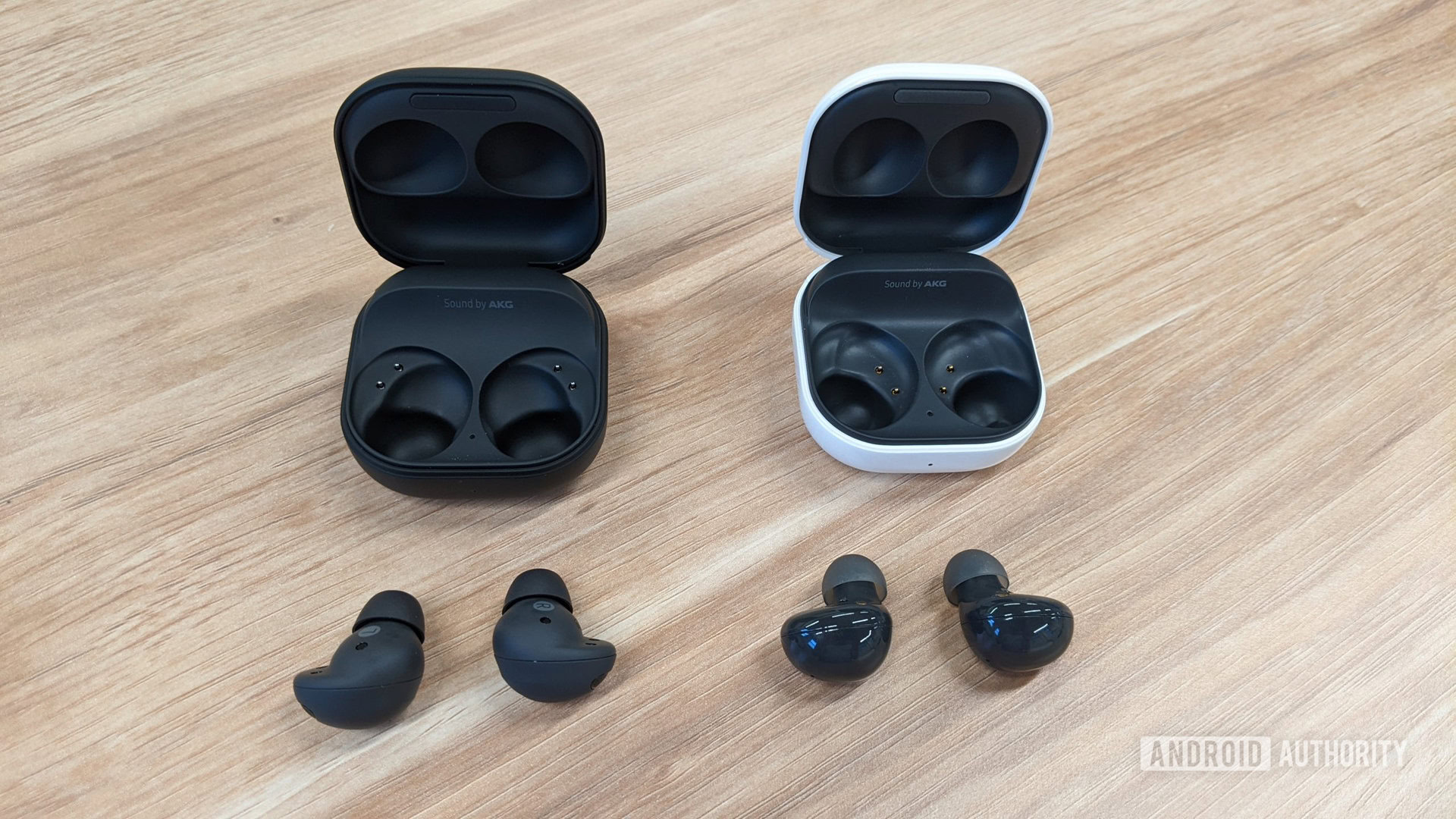 Los Samsung Galaxy Buds 2 Pro de grafito están ubicados en el exterior de la carcasa a la izquierda de los Galaxy Buds 2 de grafito fuera de la carcasa (blanca) sobre una mesa.