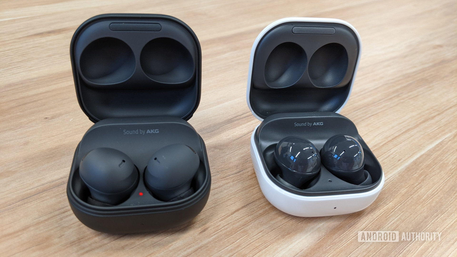 The Samsung Galaxy Buds 2 Pro in graphite sitting inside of their case to the left of the Galaxy Buds 2 in graphite inside of their (white) case on a table.