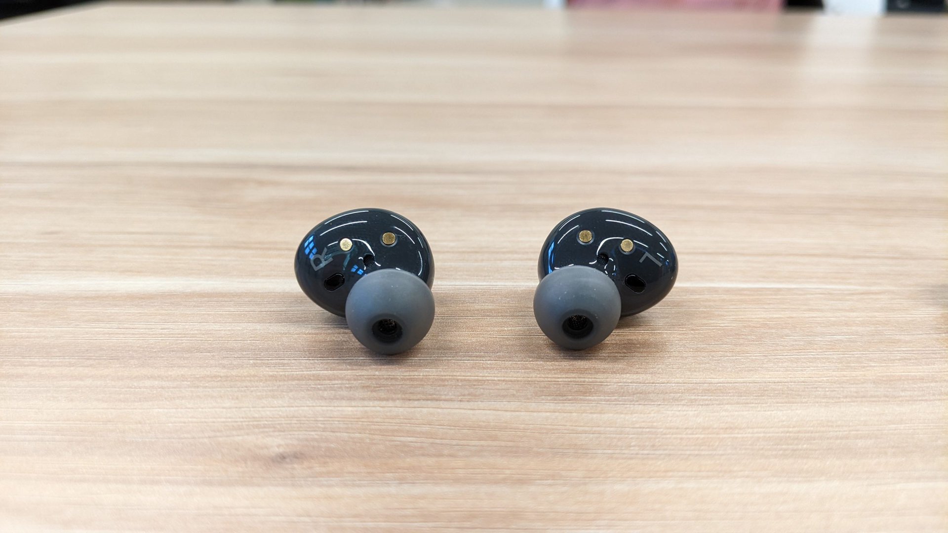The Samsung Galaxy Buds 2 in graphite outside of the case seen from the back resting on a wooden table.