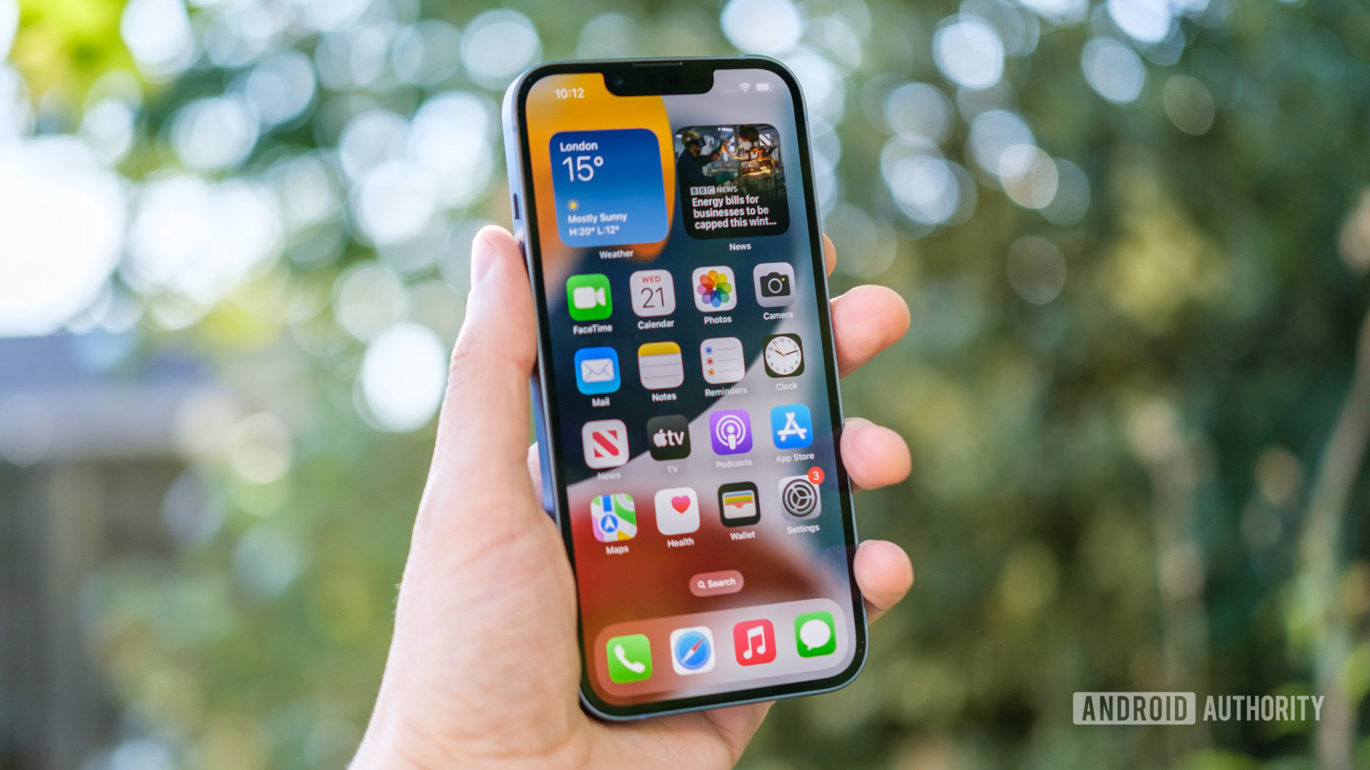 A hand holding an Apple iPhone 14 showing its homescreen in front of a blurry background of lants and leaves.