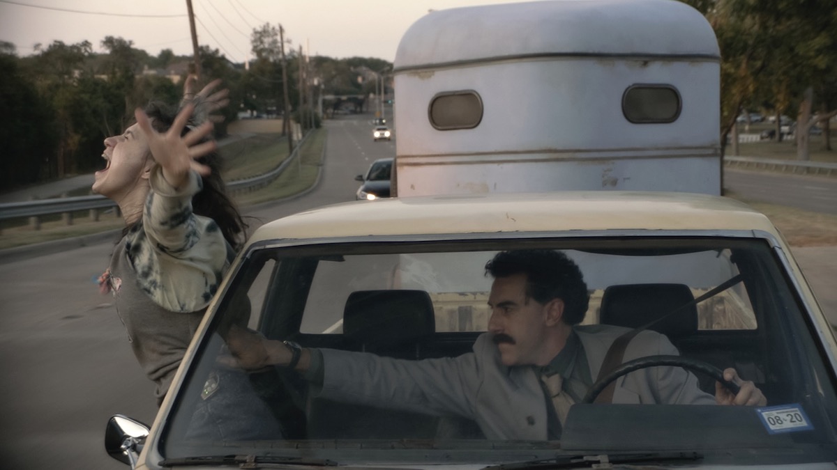 Borat pulls his daughter back into the car he's driving as she stands out the window in Borat Subsequent Moviefilm