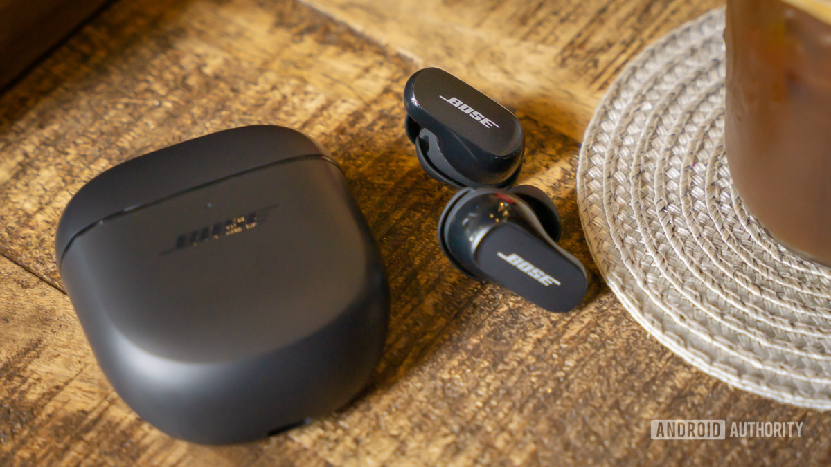 The Bose QuietComfort Earbuds II lying outside and next to their case on a wood surface beside a mug on a clear plastic coaster.