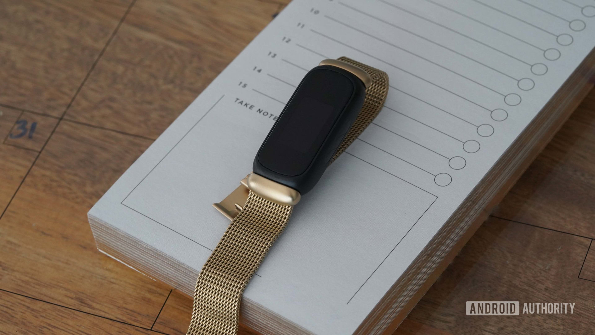 Un Fitbit Inspire 3 avec un bracelet en maille d'acier inoxydable doré repose sur un bloc-notes.