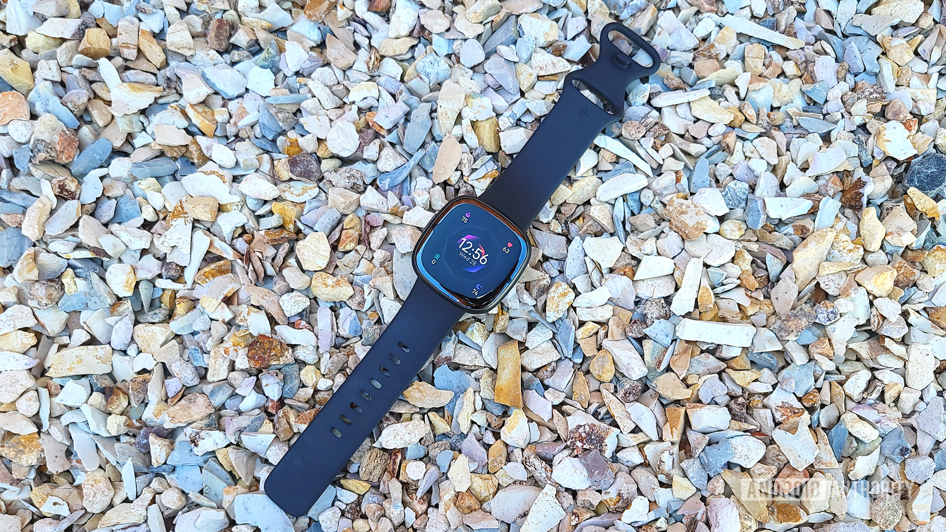 A Fitbit Sense 2 rests on pebbles, display a watch face.